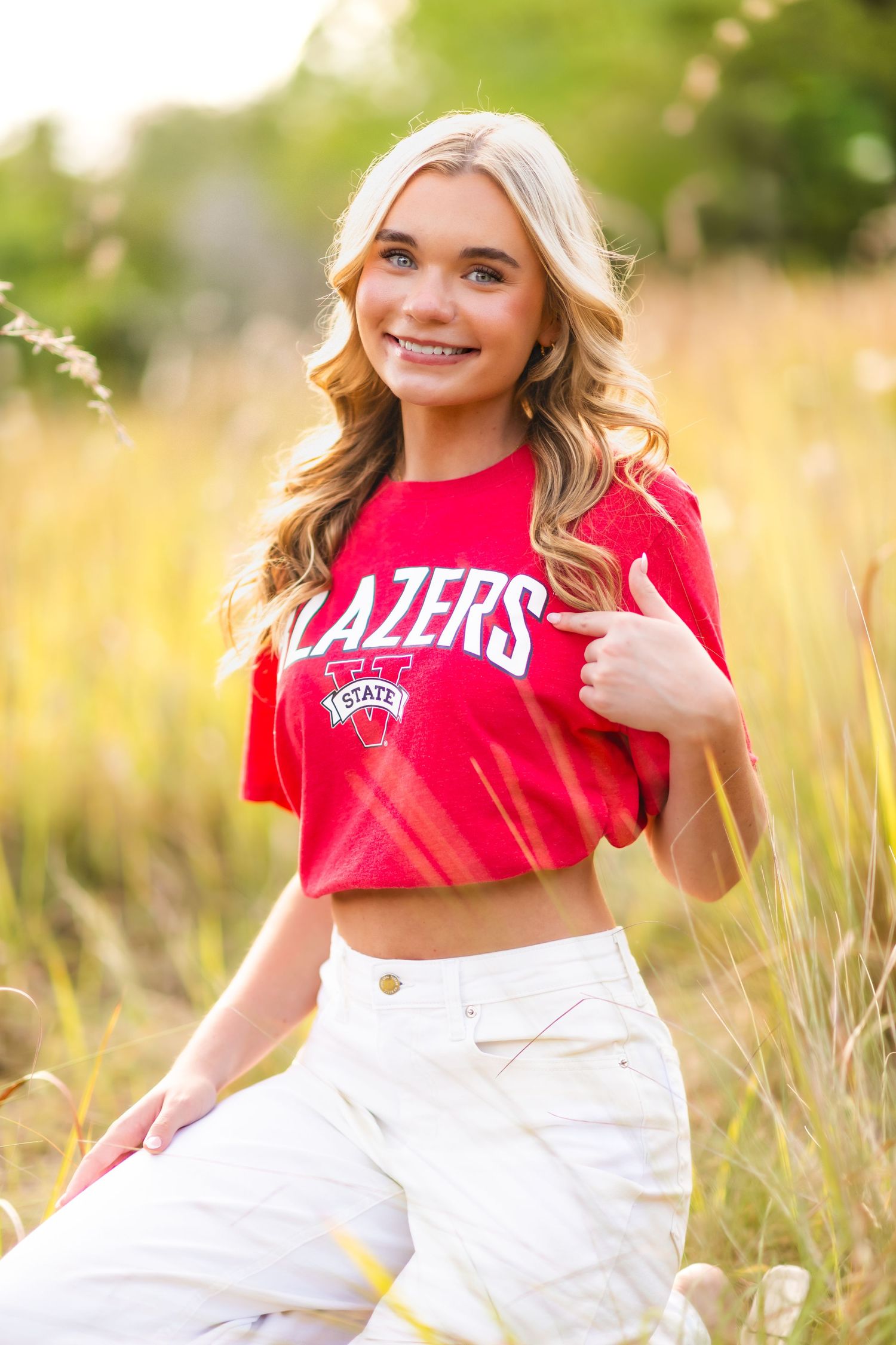 Young blonde woman smiling in a red Blazers State crop top and white jeans, posing in a golden grass field.
