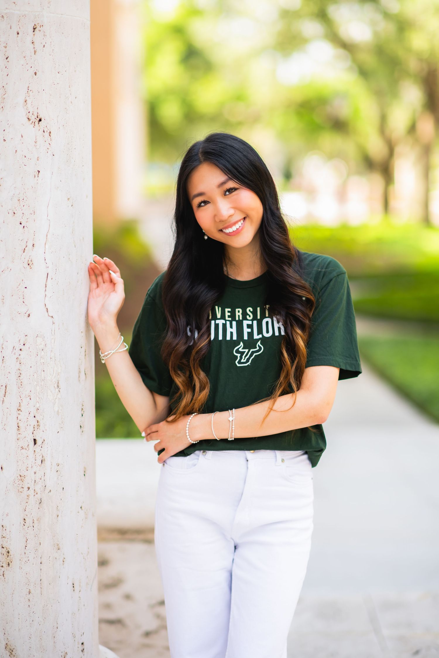 Smiling young woman in University of South Florida green t-shirt and white jeans posing outdoors on campus.