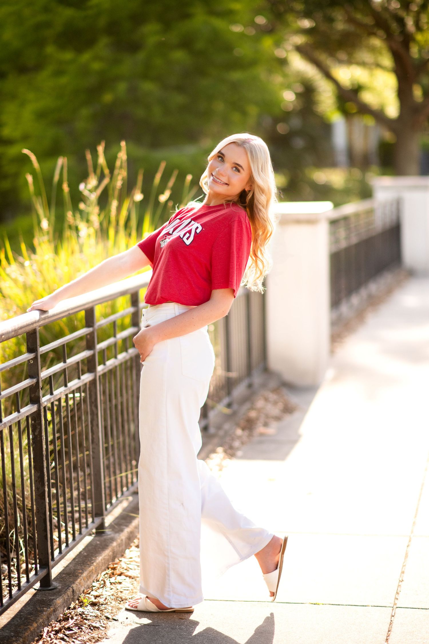 Young blonde woman in red top and white wide-leg pants posing by a railing on a sunny outdoor walkway.