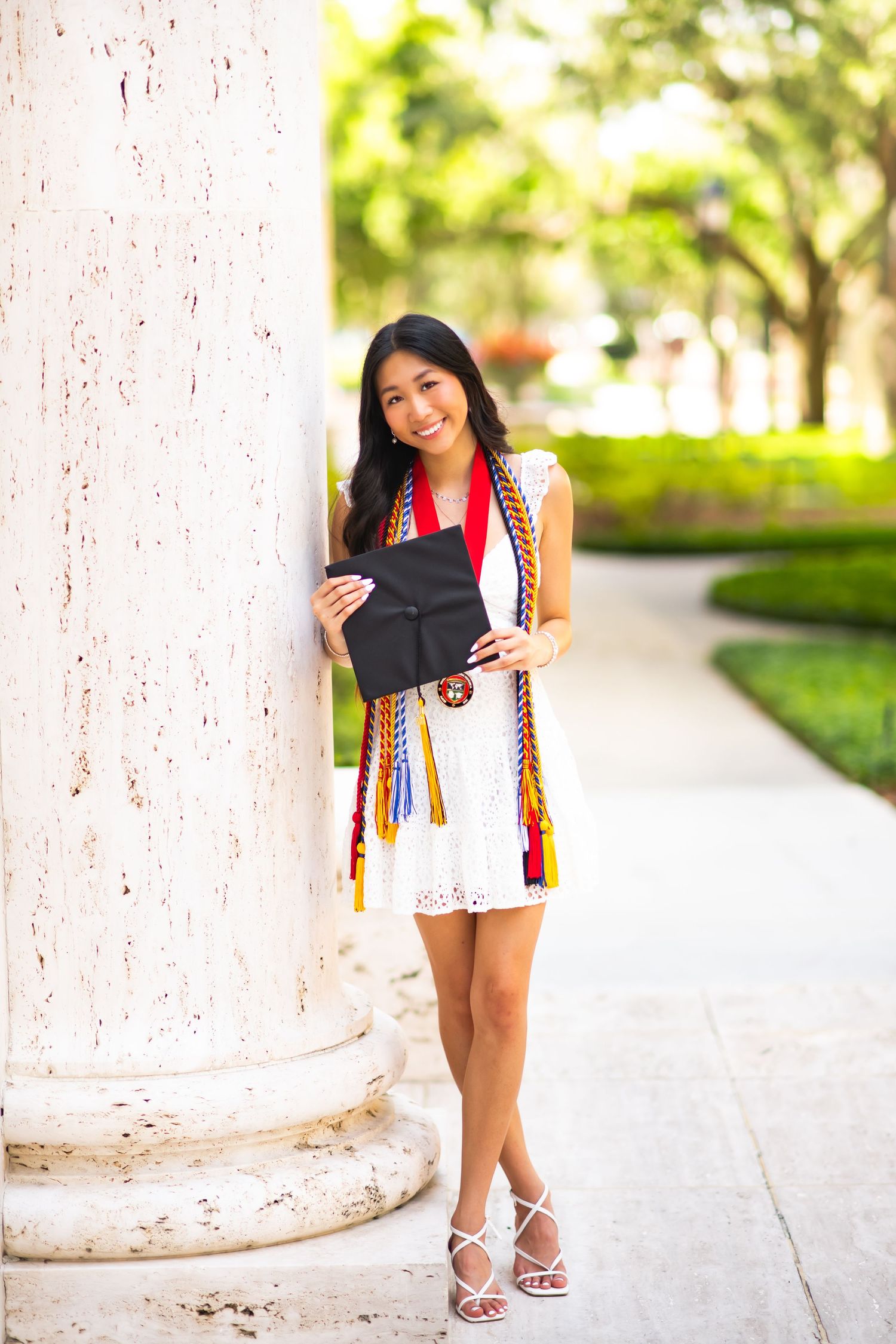 Young Asian woman in white dress holding graduation cap with colorful honor cords, posing by white column outdoors.