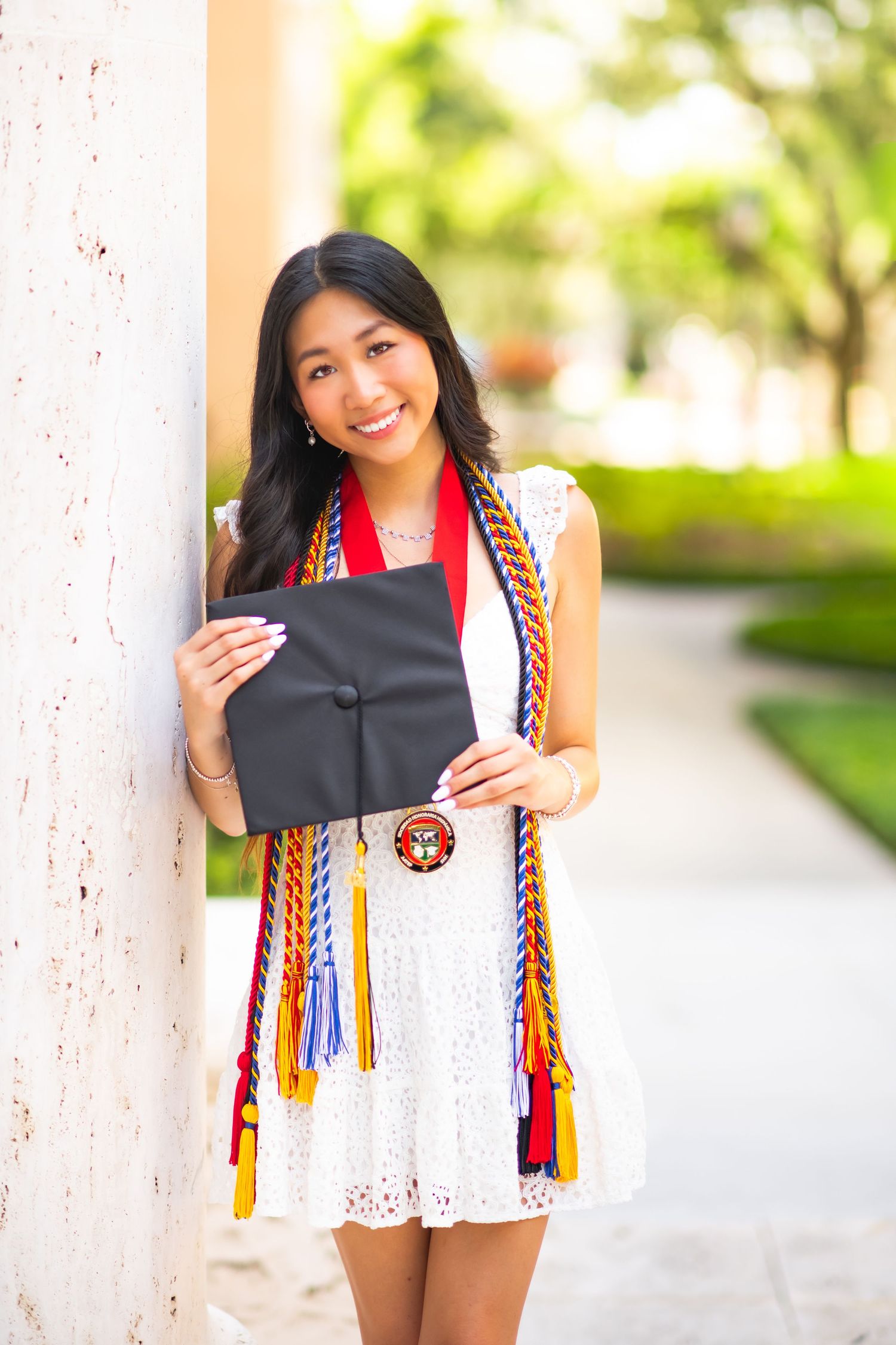 Smiling young woman in white dress holding graduation cap with colorful honor cords outdoors.