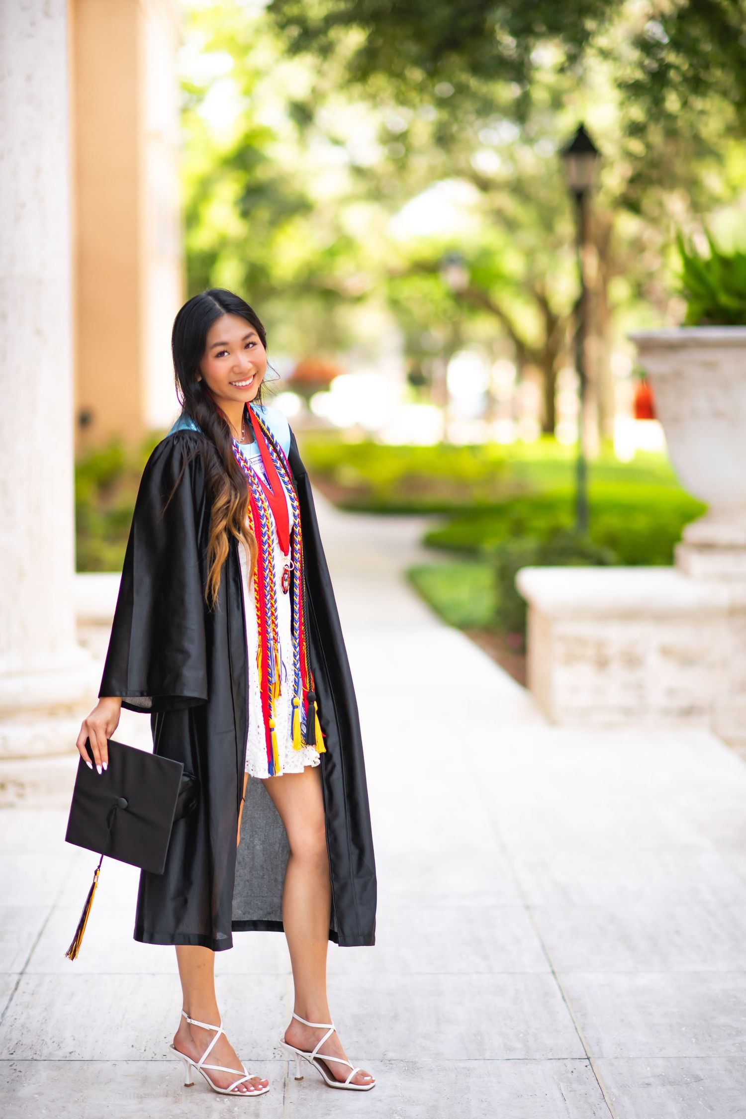 Young woman in black graduation gown and honor cords smiles while holding cap outdoors on a sunny campus.