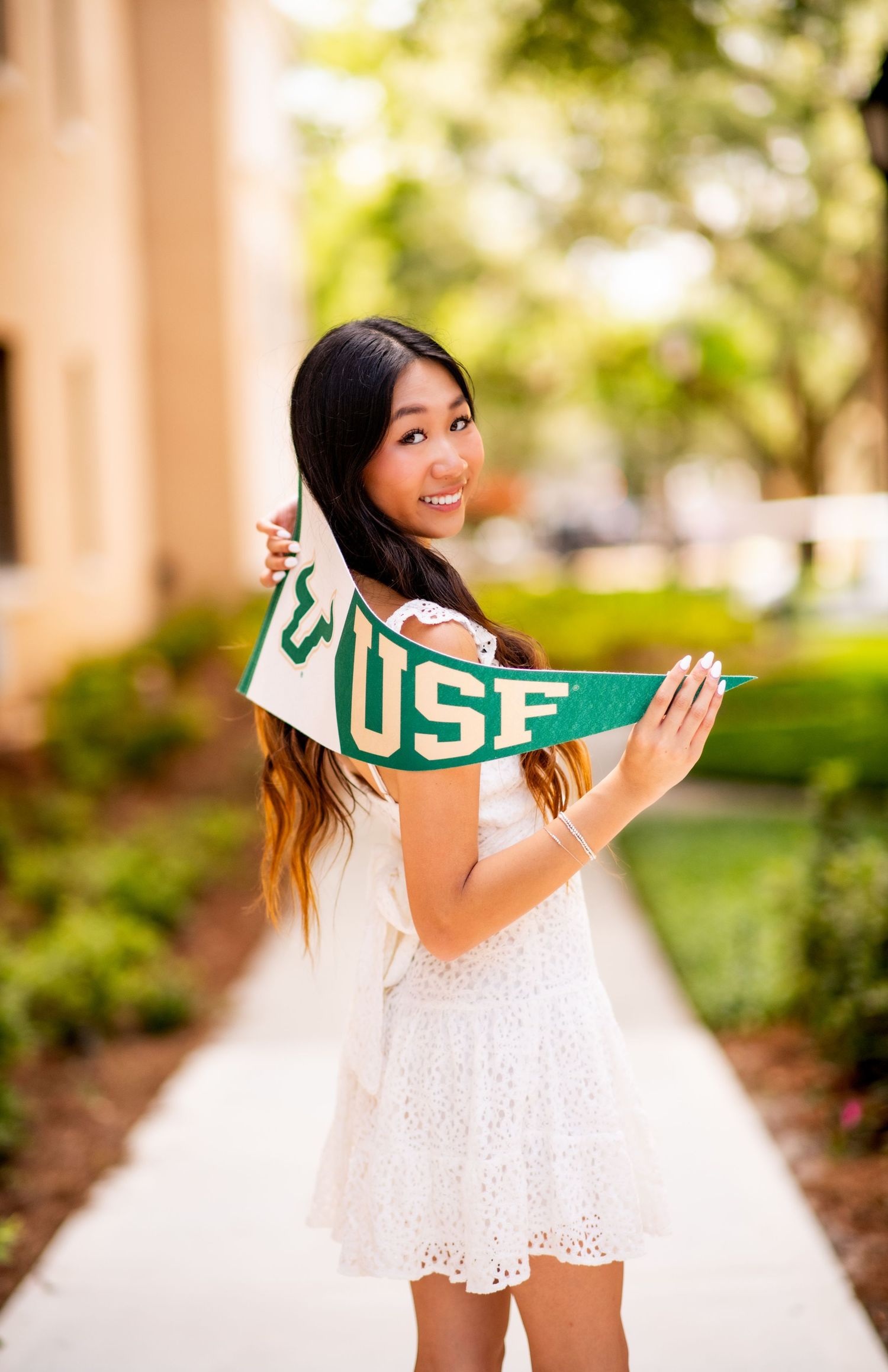 A smiling young woman in a white dress holds a green USF pennant flag on a sunny campus walkway.