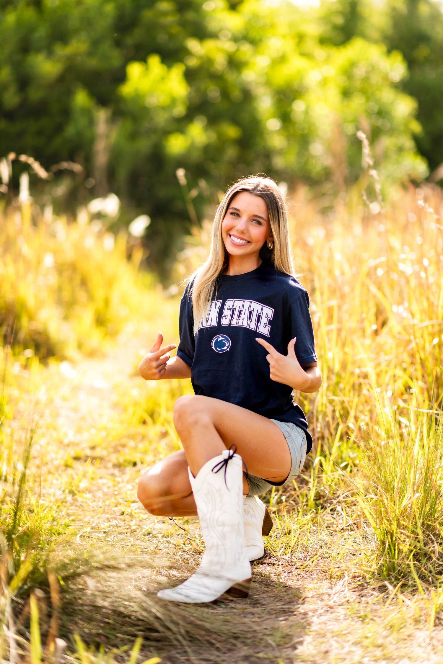 Smiling blonde woman in Penn State shirt, denim shorts, and white cowboy boots posing outdoors in golden grass field.