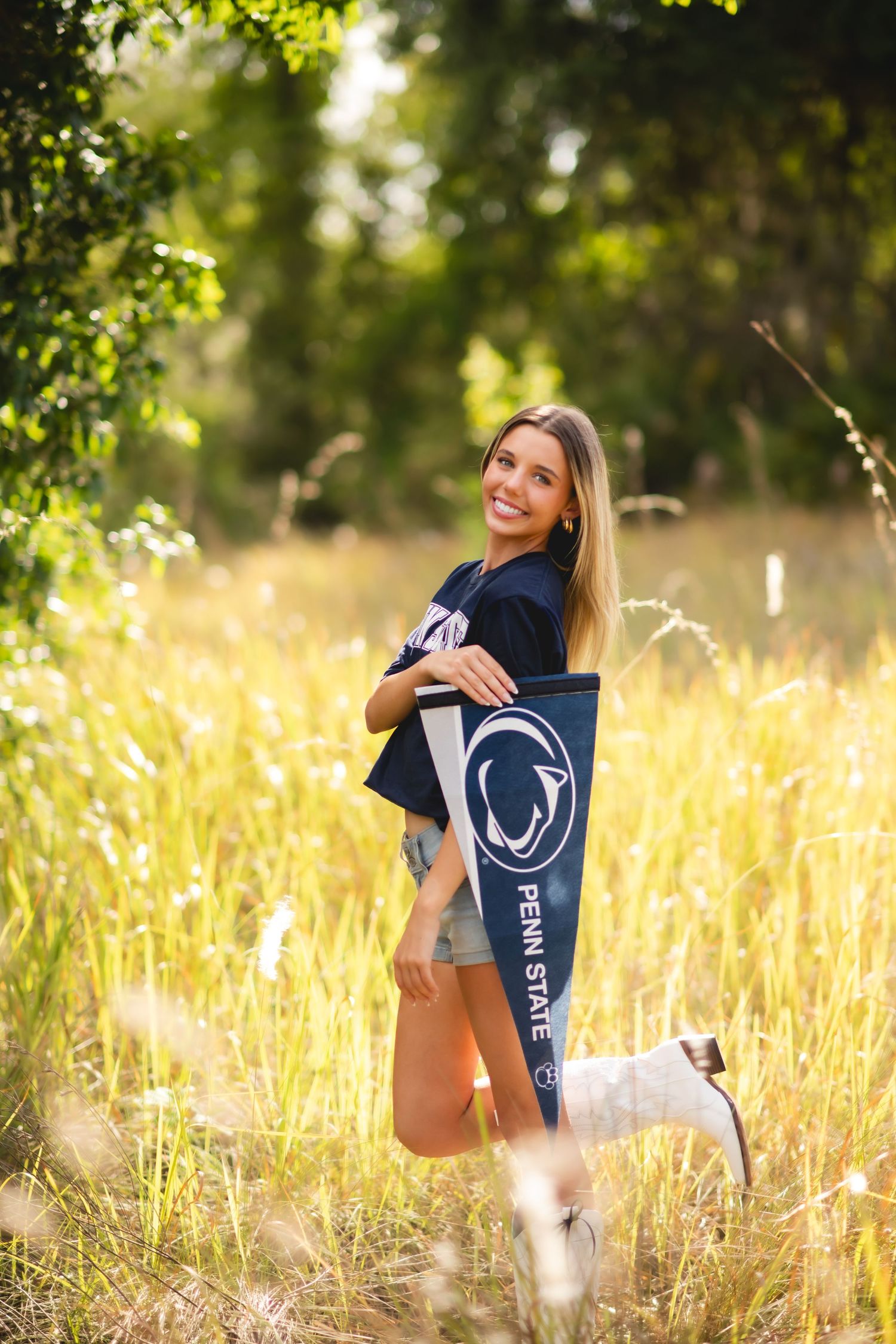 Smiling young woman holding a Penn State pennant and wearing a Penn State shirt, posing in a sunny golden field.