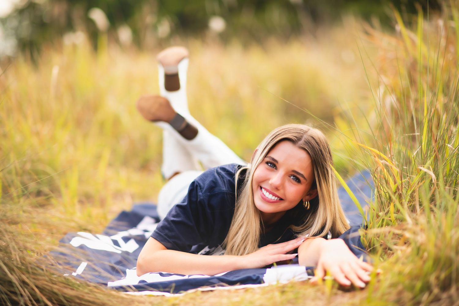 Smiling blonde woman lying on a blanket in a golden grass field, wearing a navy outfit, posing for a portrait photo.