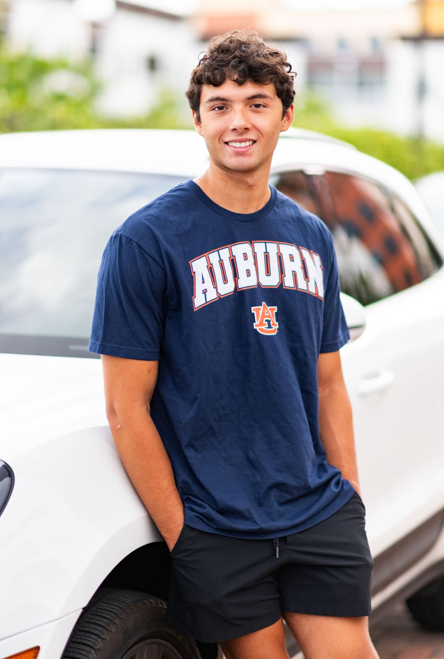 Young man smiling in navy Auburn University t-shirt and black shorts, leaning against a white car outdoors.
