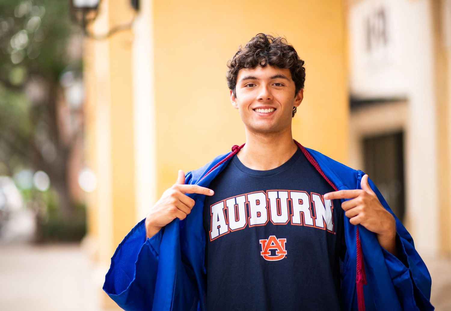 Smiling young man proudly pointing to his Auburn University navy blue t-shirt while opening a blue jacket outdoors.