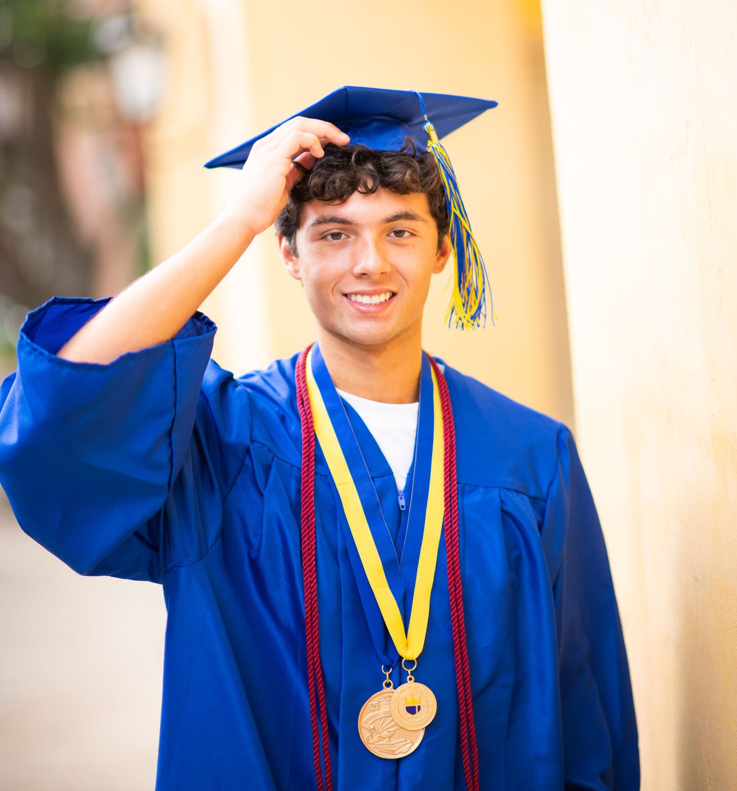 Smiling young man in blue graduation cap and gown wearing a gold medal, posing outdoors for senior photos.