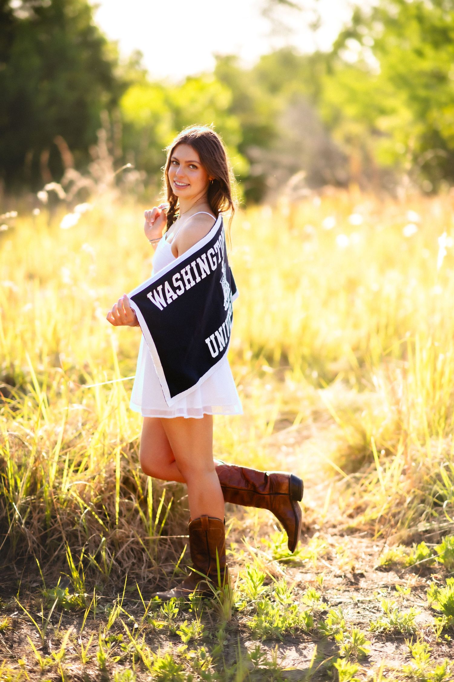 Young woman in white dress and cowboy boots holding Washington University banner in golden field.