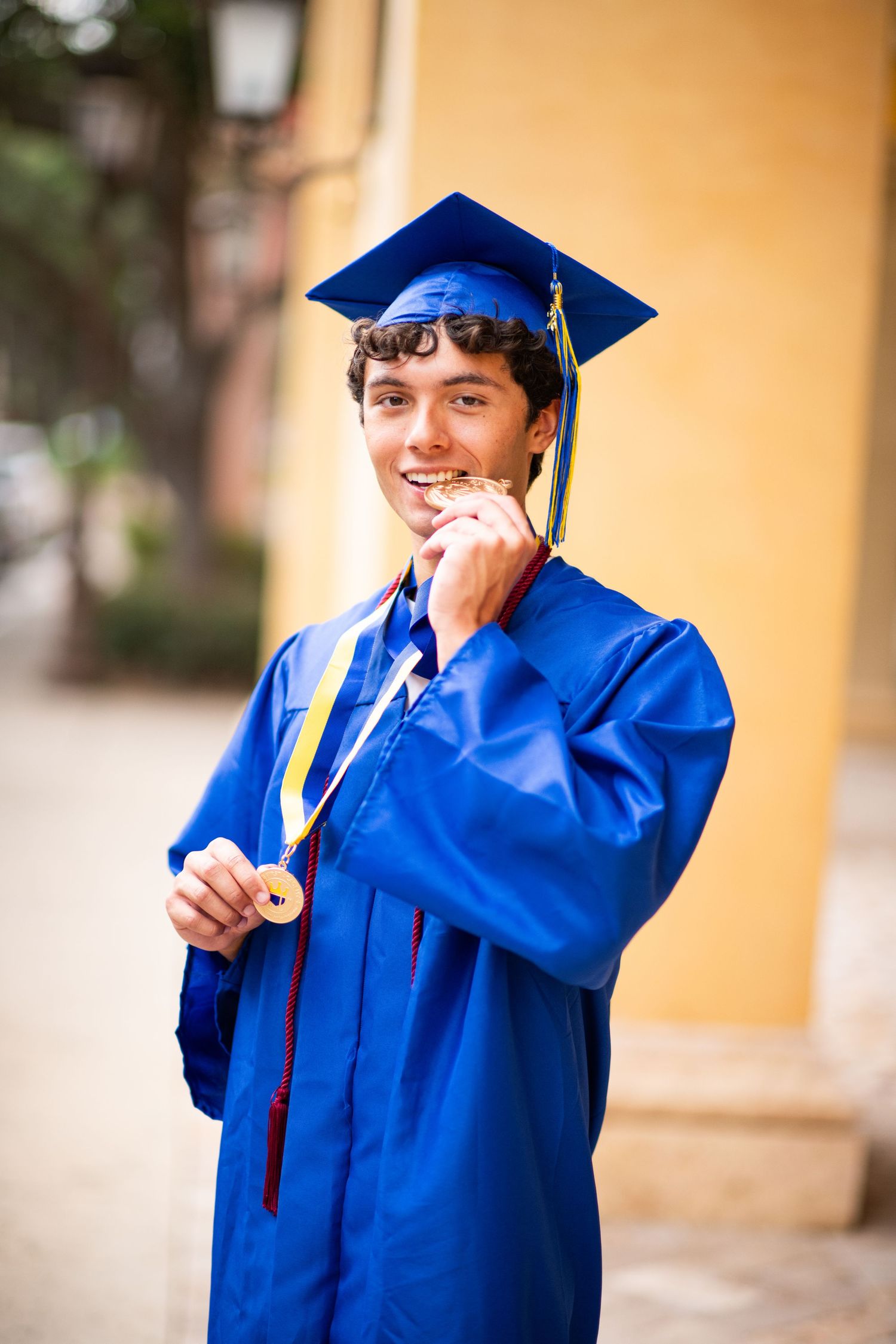 Smiling young man in blue graduation cap and gown proudly displaying a gold medal outdoors.