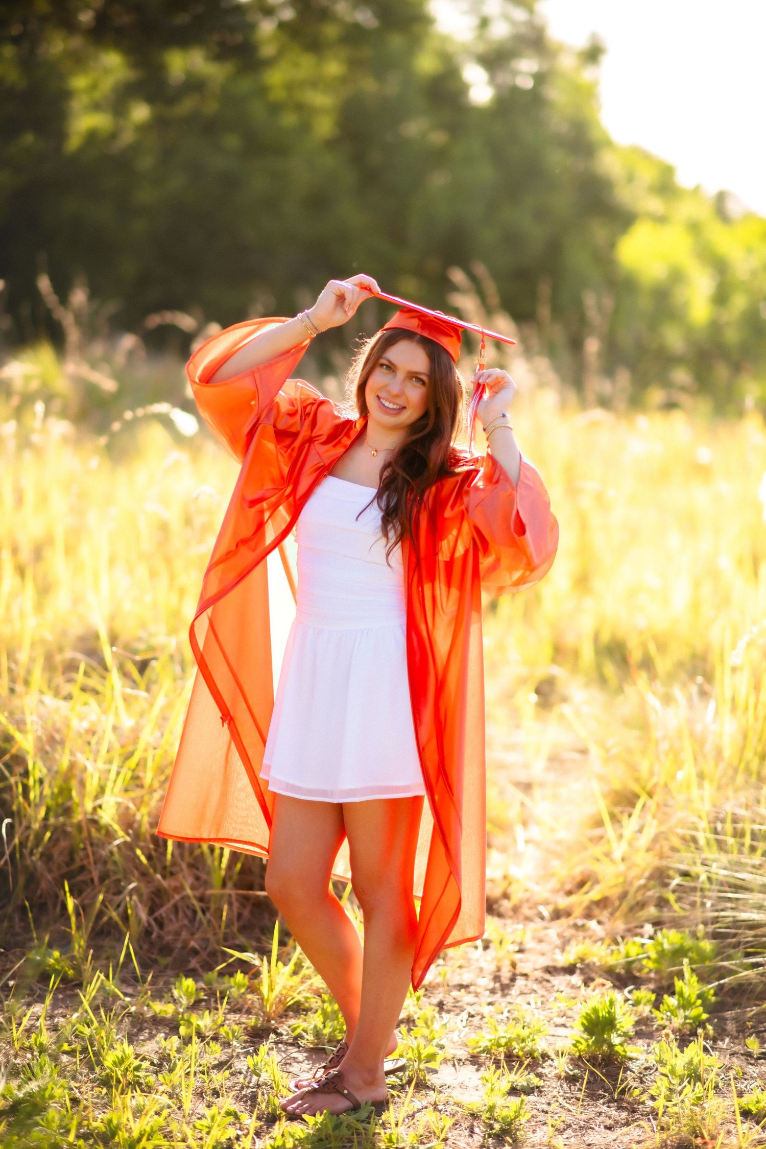 Young woman in white dress and red graduation gown holds cap overhead in sunny field.