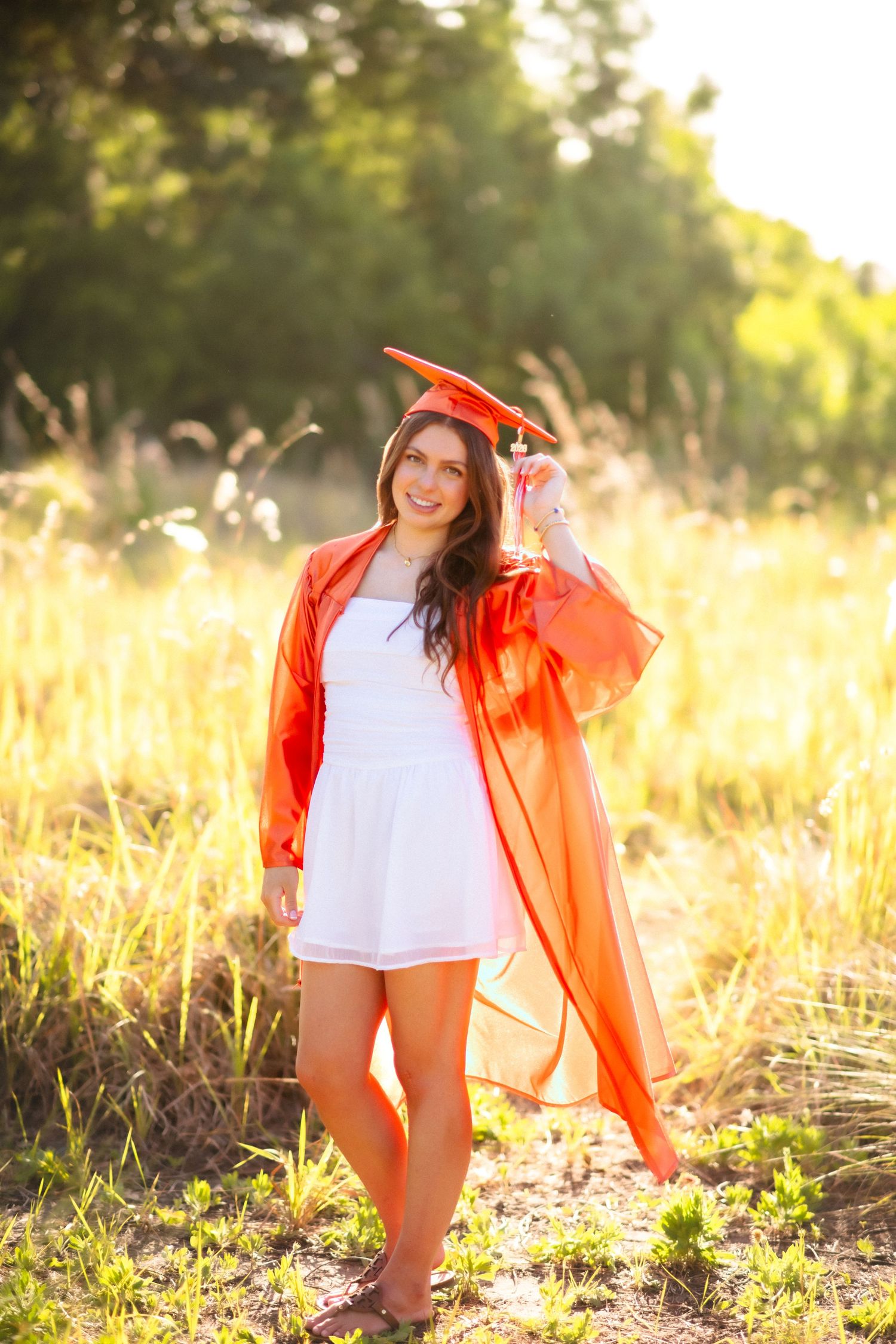 Young woman in orange graduation cap and gown over white dress poses in sunny field.