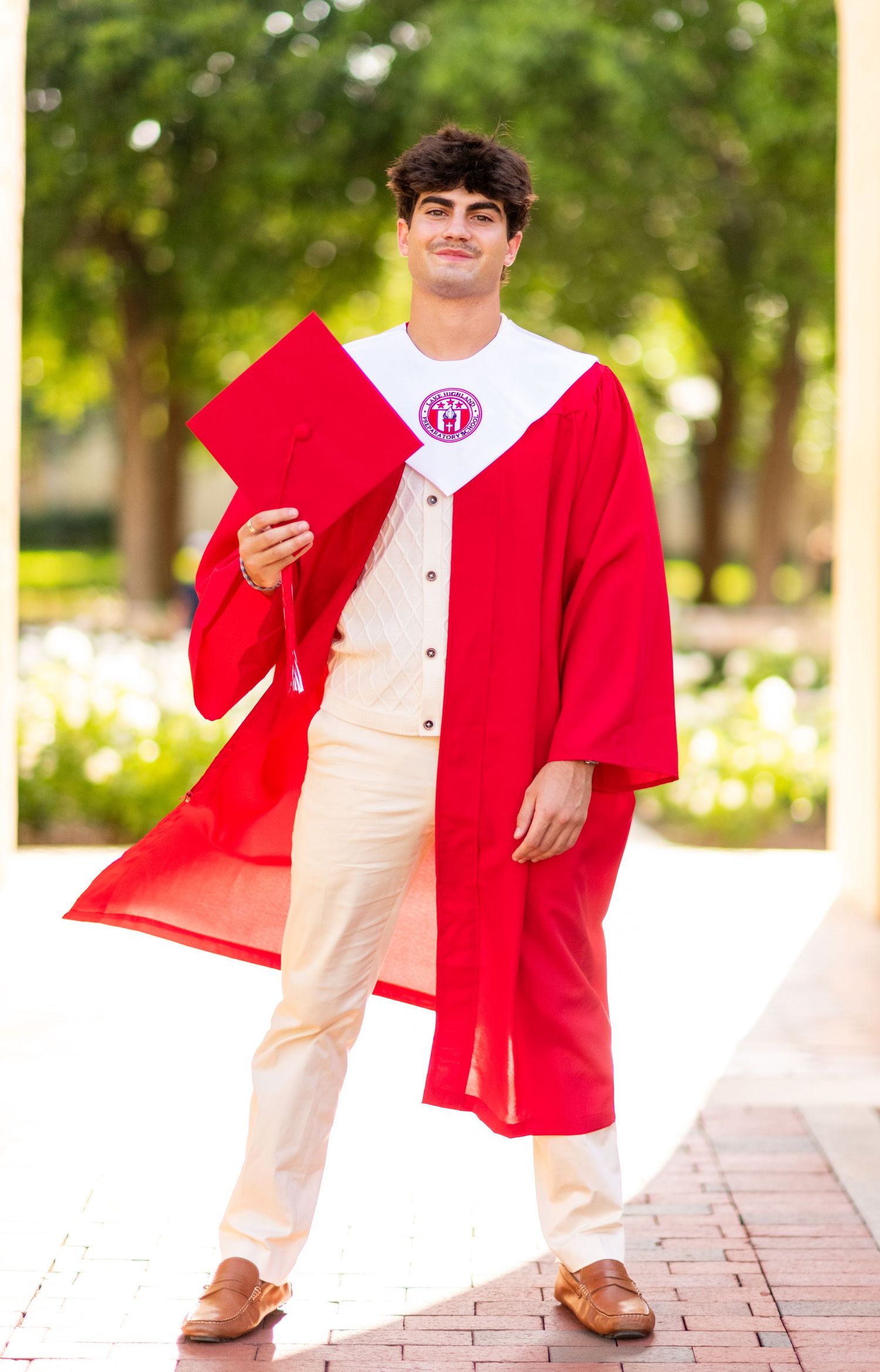 Young man in red graduation gown and cap smiling outdoors on a brick path with green trees in background.