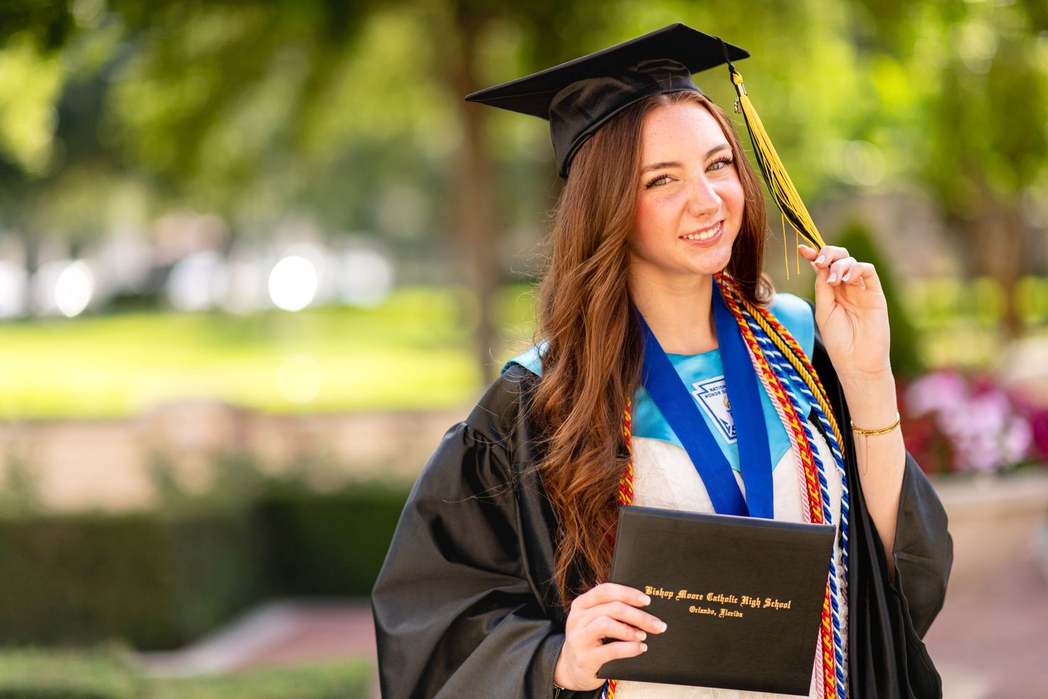 Smiling female graduate in black cap and gown holding diploma outdoors, wearing colorful honor cords and blue medal.