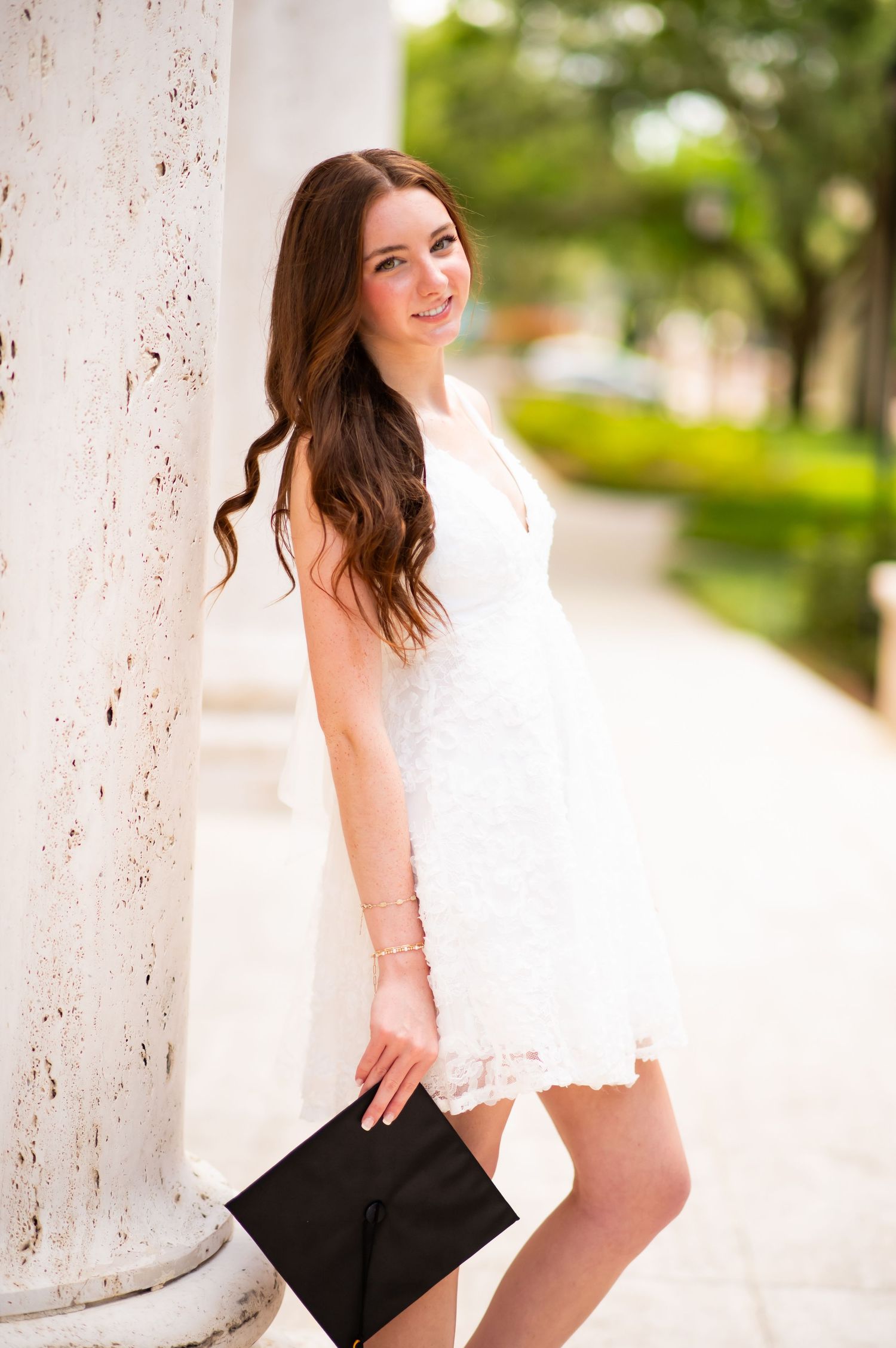 Young woman in white lace dress holding black clutch, leaning against white column outdoors.