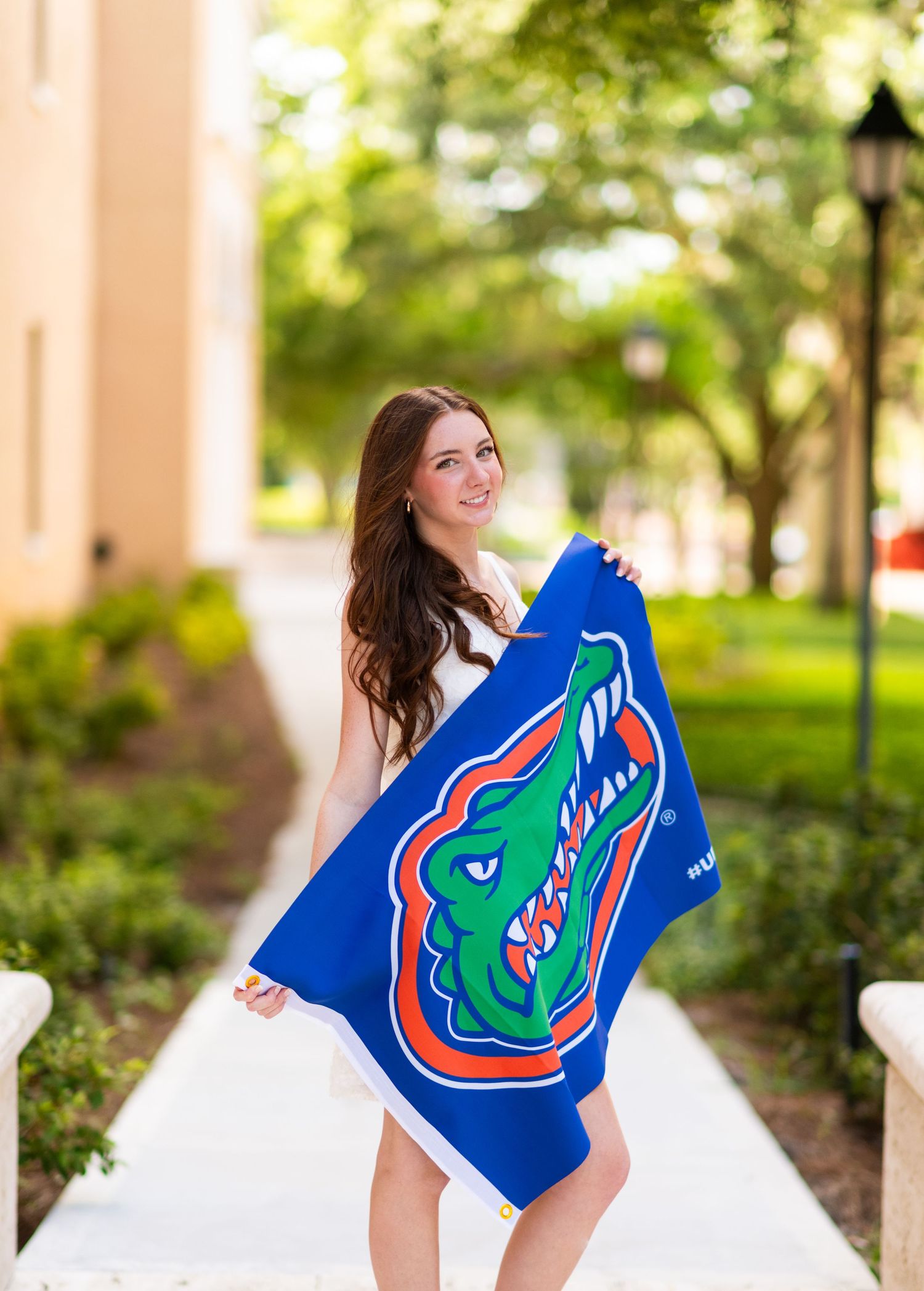 Young woman in white dress holding a University of Florida Gators flag outdoors on a sunny campus pathway.