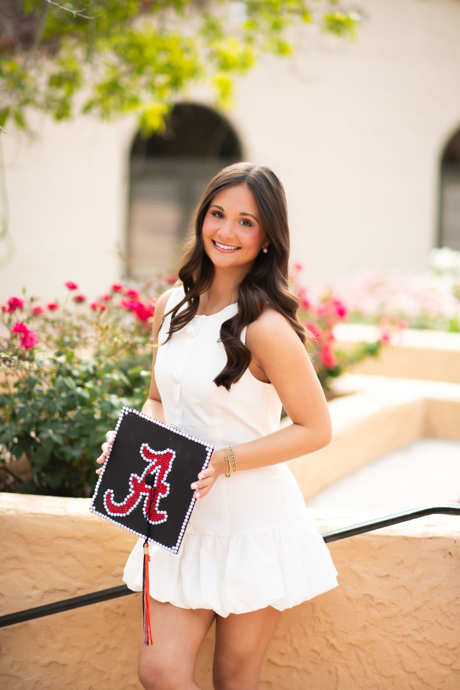 Young woman in white dress holding decorated University of Alabama graduation cap outdoors near pink flowers.