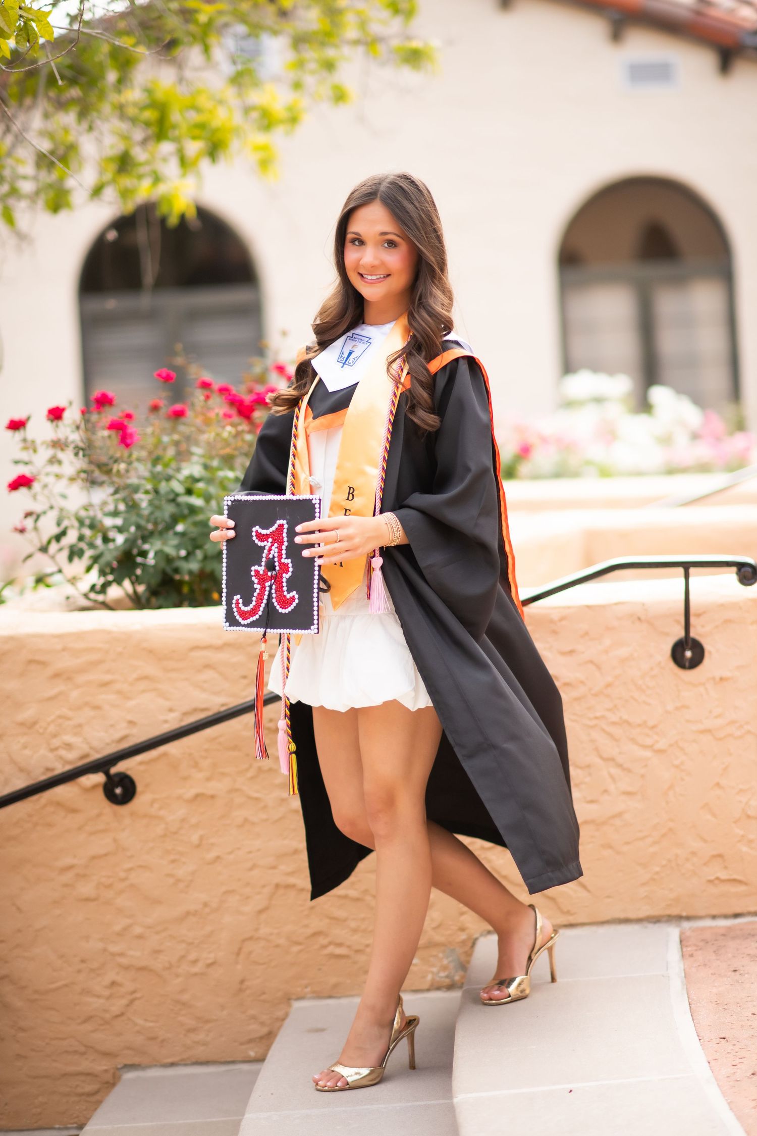 Young woman in black graduation gown holding University of Alabama cap on outdoor steps.