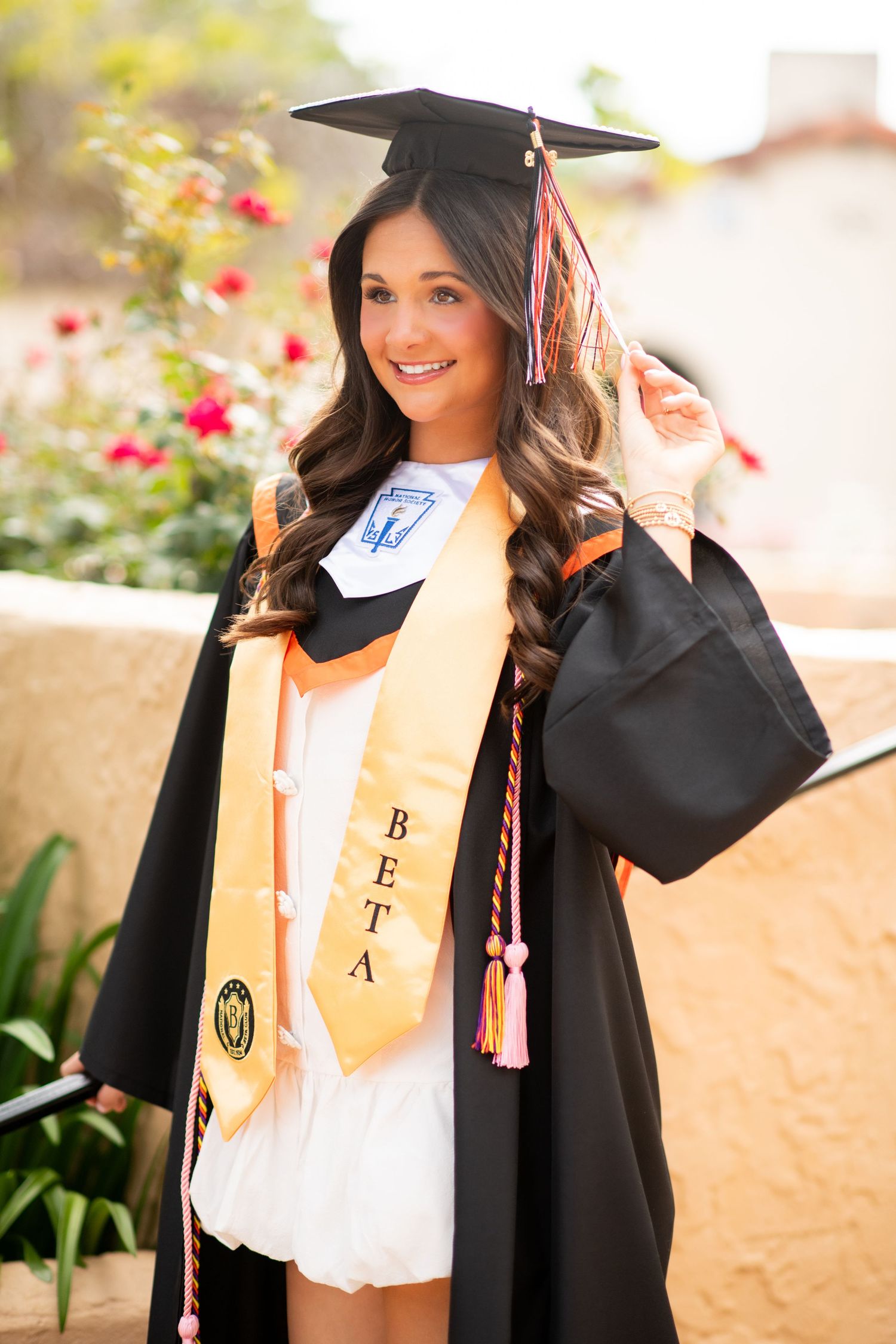 Smiling female graduate in black cap and gown with gold Beta honor stole posing outdoors at graduation ceremony.