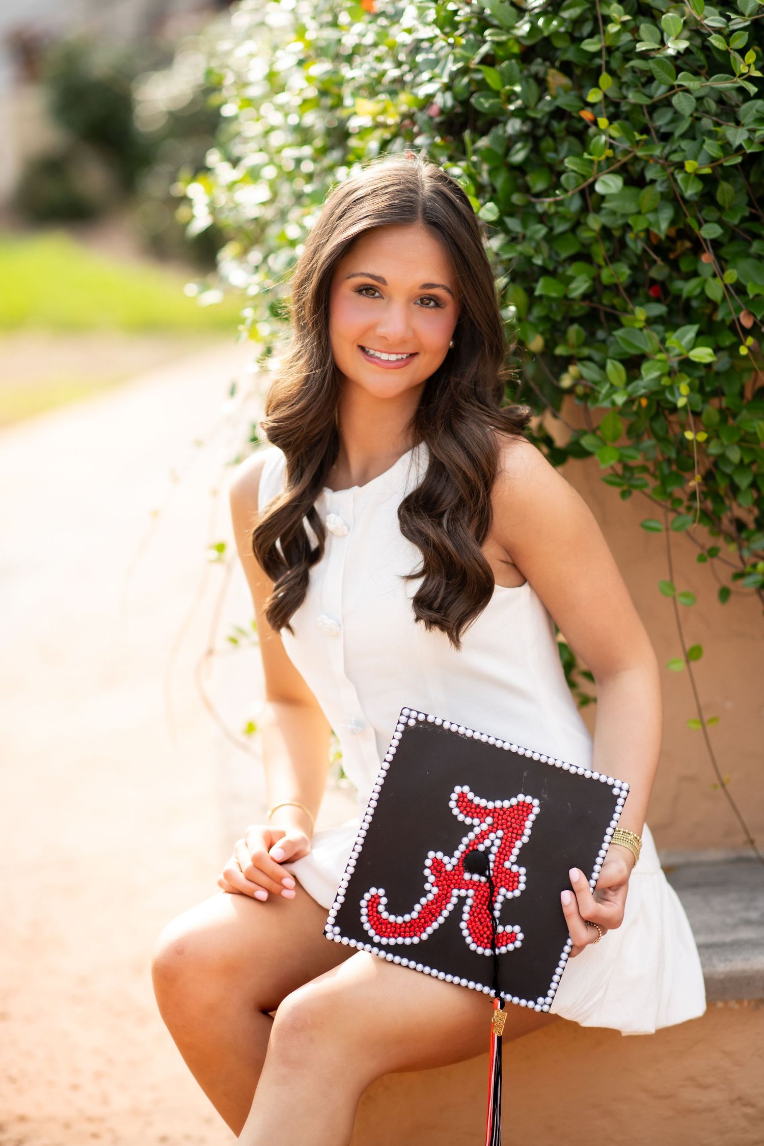 Smiling young woman in white dress holding decorated University of Alabama graduation cap outdoors.