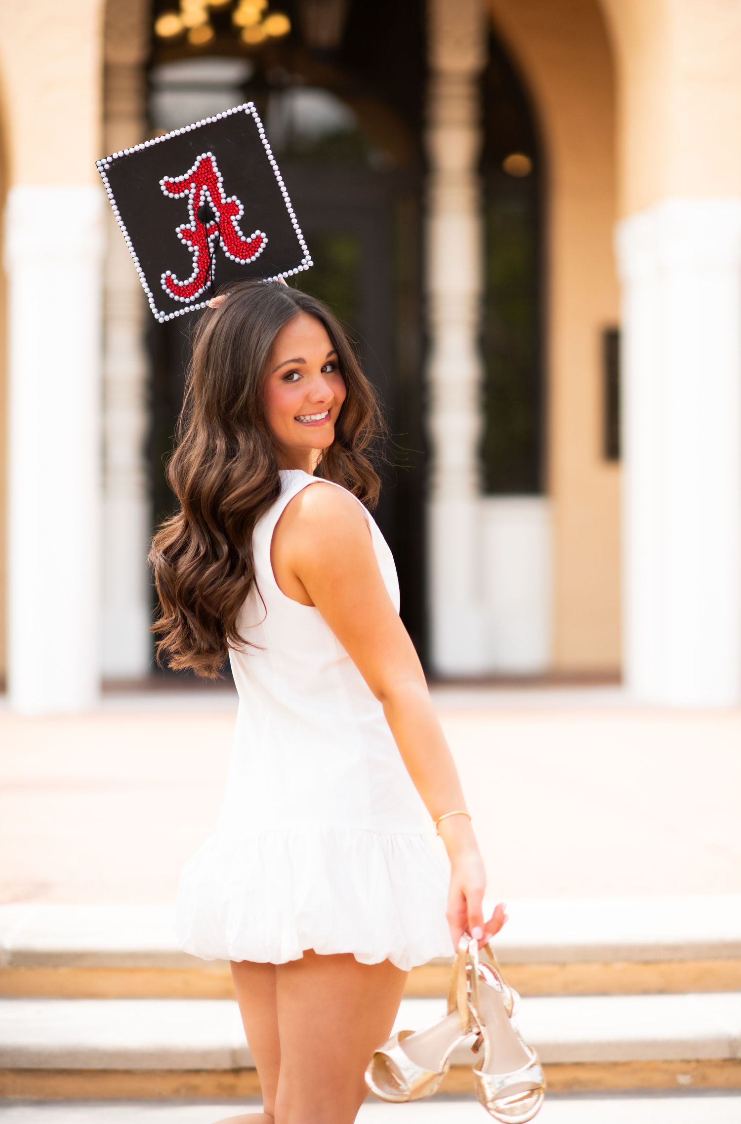 Smiling woman in white dress holding heels, posing with University of Alabama graduation cap outdoors.