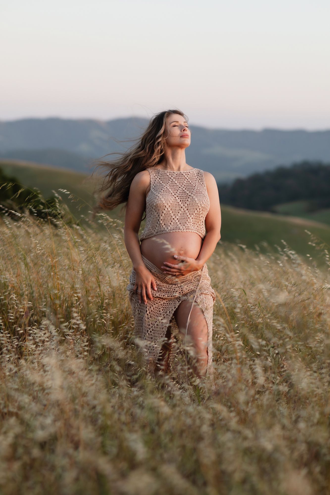 Expectant mother in tan crochet outfit closes eyes and poses in golden field at blue hour with hills. North Conway maternity photographer