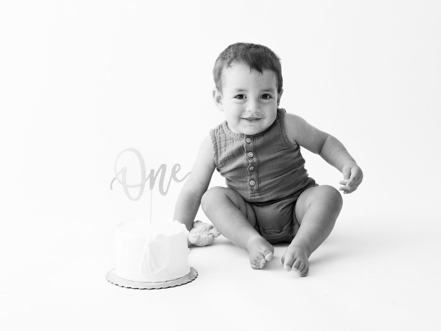 Black and white photo of a baby boy sitting beside a first birthday cake with a One topper, smiling at camera.