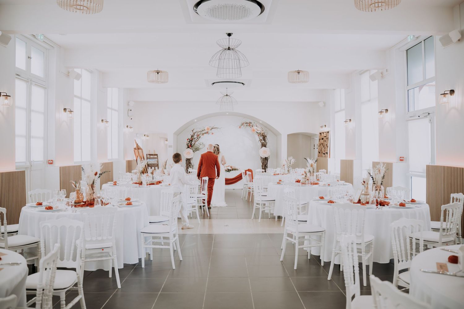 Elegant white wedding reception hall with round tables, white chairs, and a floral arch centerpiece in warm red tones.