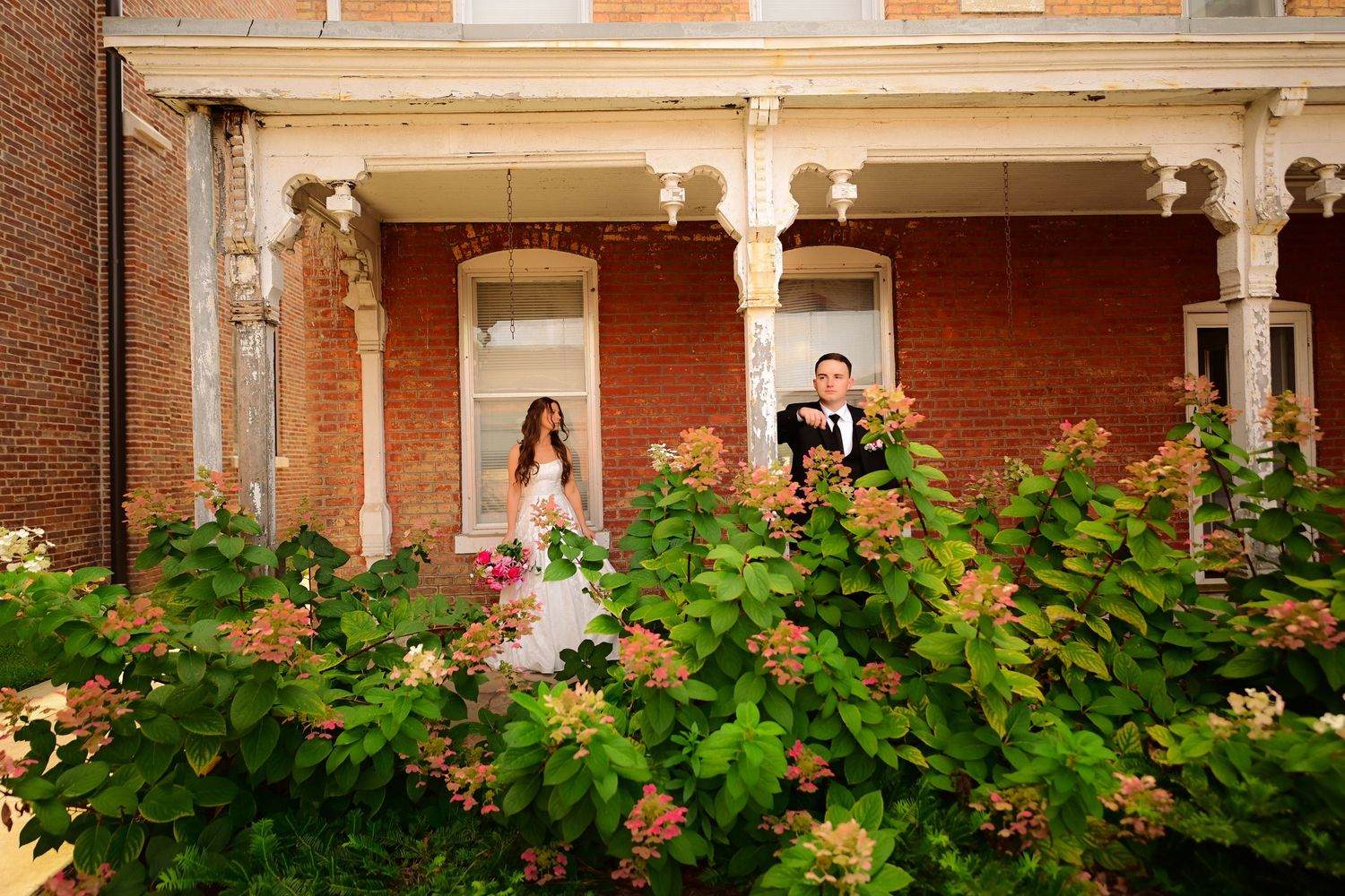 Bride and groom posing on a brick mansion porch surrounded by lush green hydrangea bushes on their wedding day.