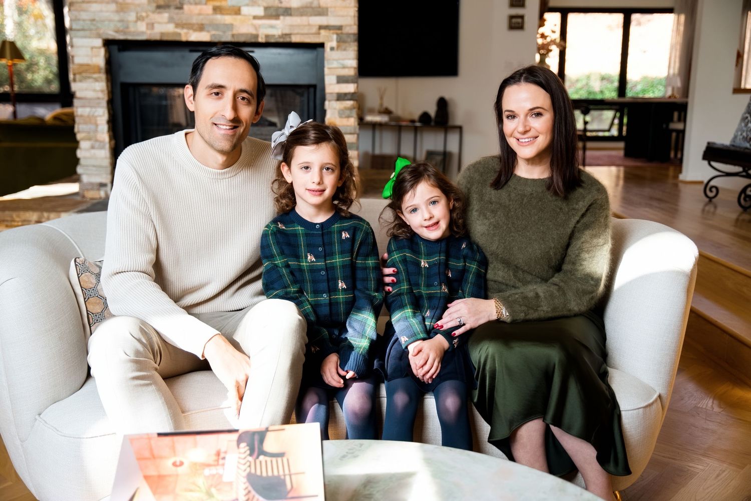 Family of four smiling together on a white sofa in a cozy living room, two young girls in matching plaid outfits.