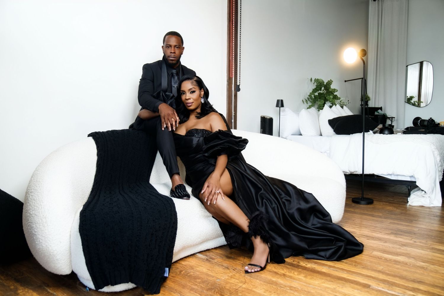 Elegant couple in matching black formal attire posing on a white curved sofa in a modern studio setting.