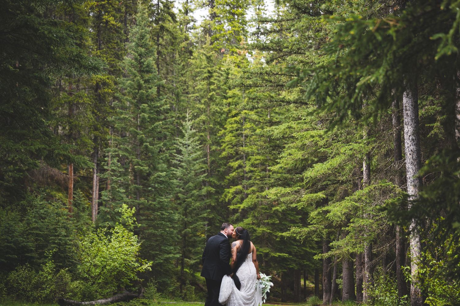 Bride and groom share a romantic moment under a white umbrella on a scenic overlook with forest and river views.
