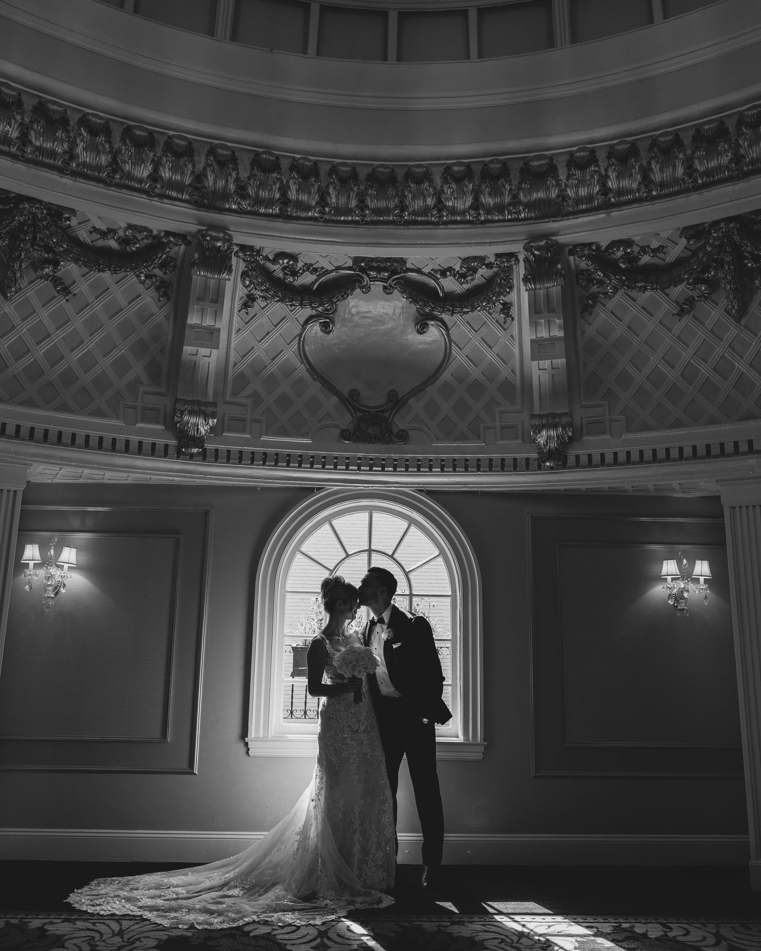 Bride and groom silhouetted against arched window in ornate ballroom, black and white wedding portrait.