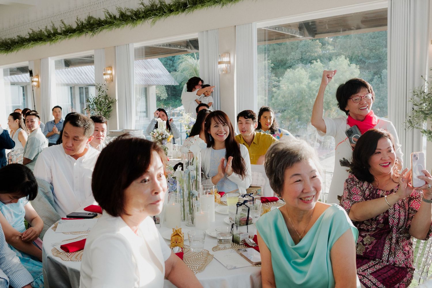 Guests laughing and celebrating at an elegant indoor wedding reception with white decor and greenery.