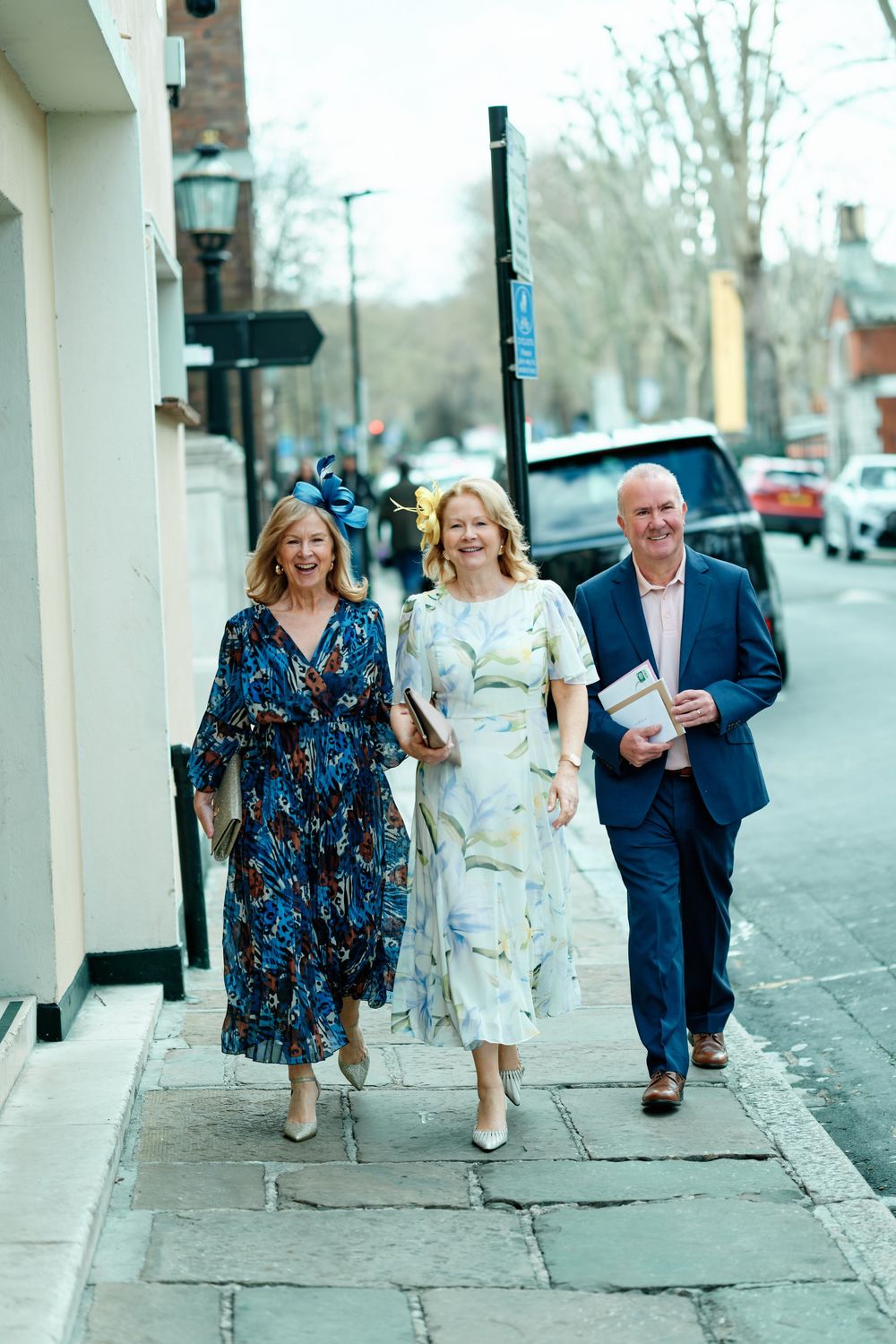 Three elegantly dressed adults walking together on a sunny sidewalk, two women in floral dresses and a man in a blue suit.