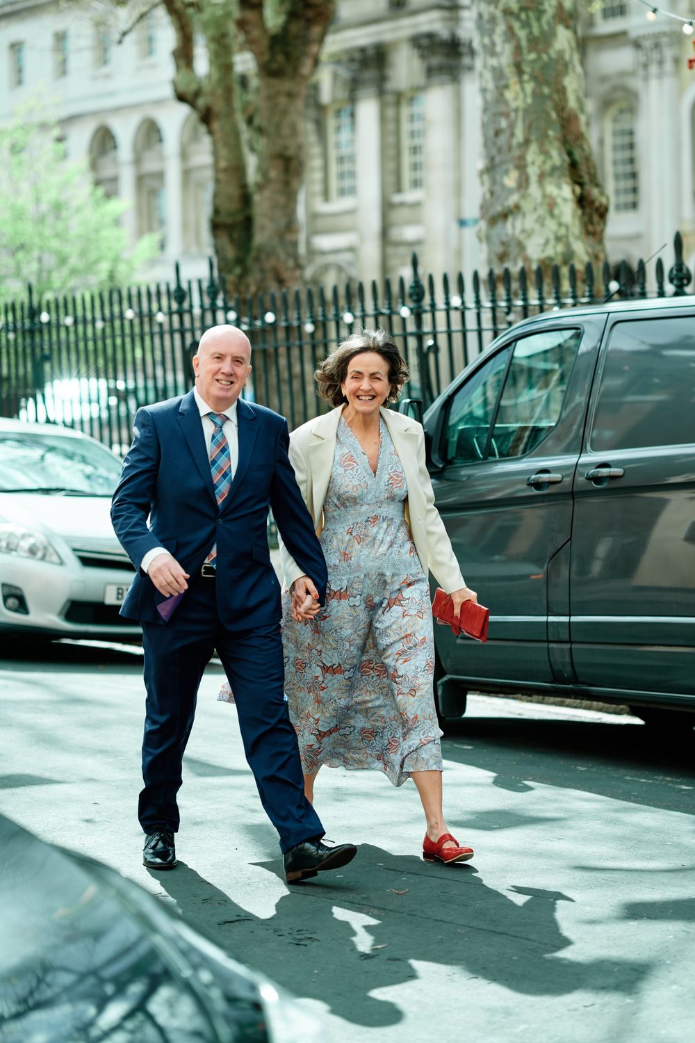 Smiling couple walking hand-in-hand across a city street, man in blue suit, woman in floral dress with cream jacket.
