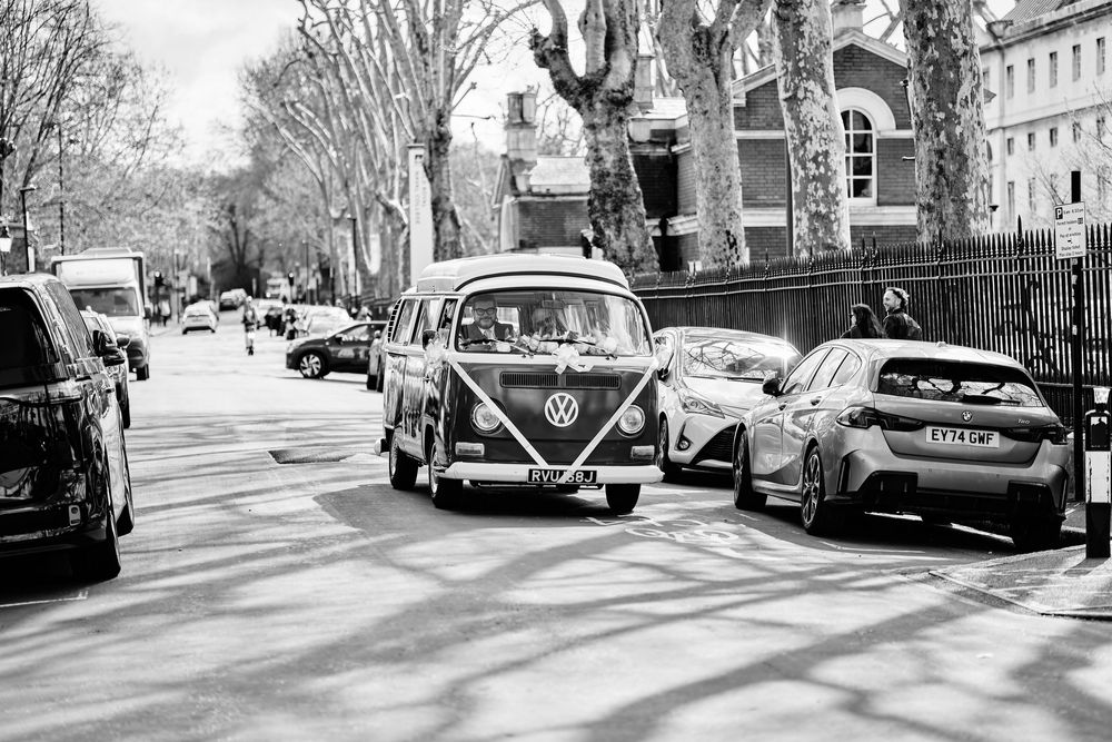 Vintage VW camper van driving down a tree-lined street in black and white urban photography.