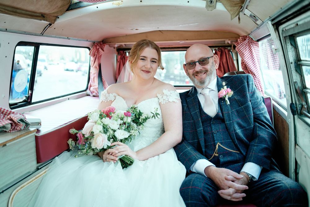Bride and groom smiling inside a vintage VW campervan, bride holding a pink and white bouquet on their wedding day.