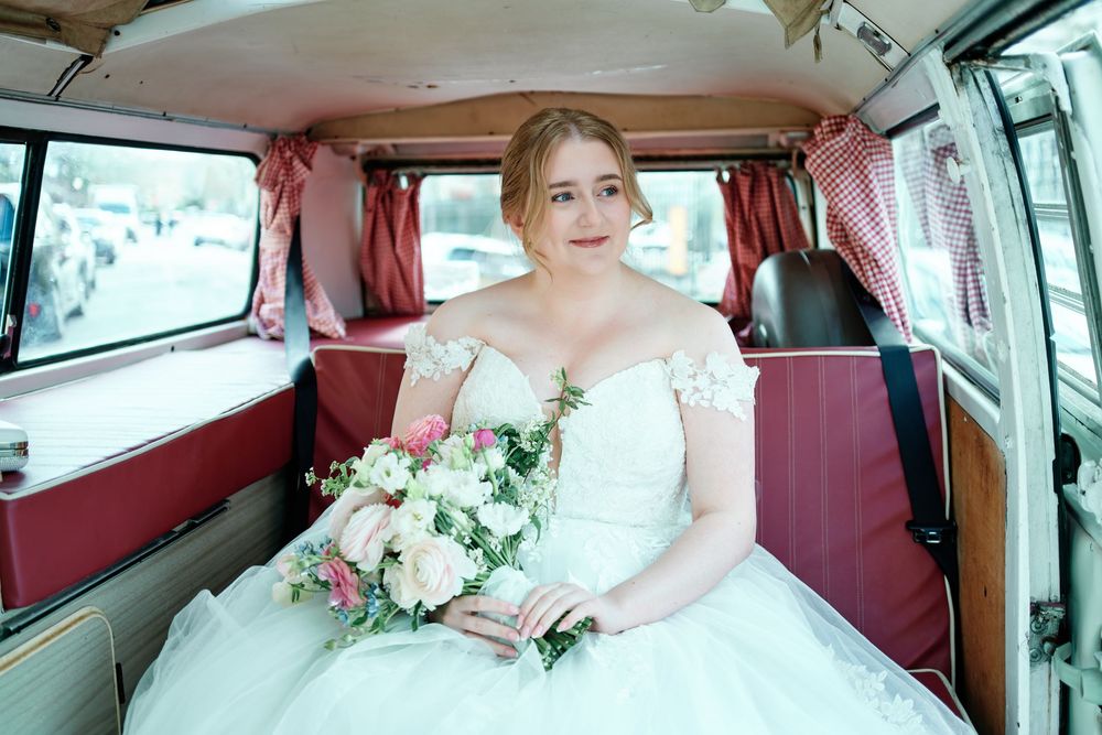 Bride in white gown holding pink bouquet seated inside vintage red and white VW camper van.