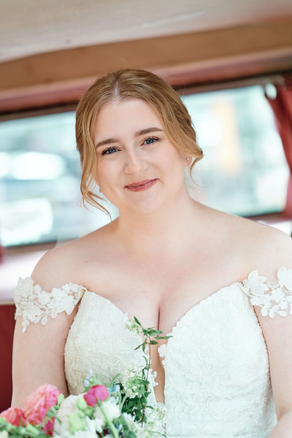 Smiling bride in off-shoulder lace wedding gown holding pink and white bouquet, seated inside a vintage vehicle.