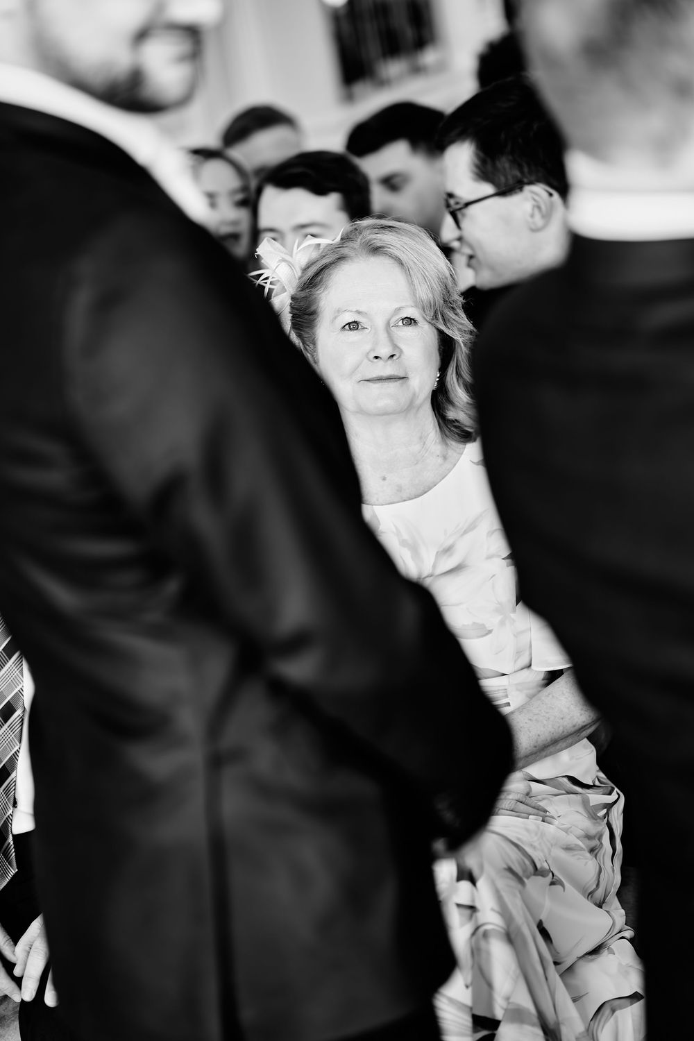 Black and white photo of an elegant older woman in formal attire surrounded by suited guests at a wedding ceremony.