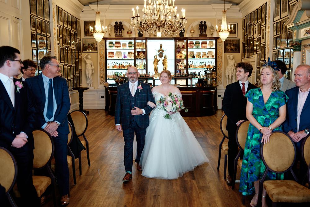 Bride walks down the aisle in a white gown at an elegant indoor wedding ceremony with wooden floors.