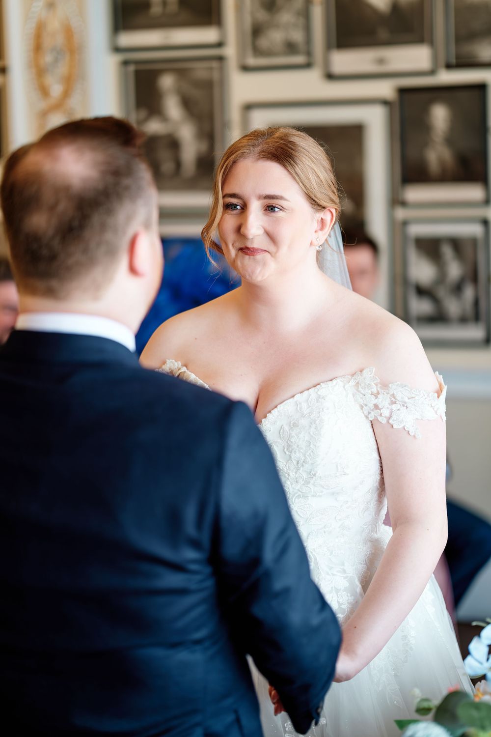 Bride in lace off-shoulder gown holding hands with groom during indoor wedding ceremony.