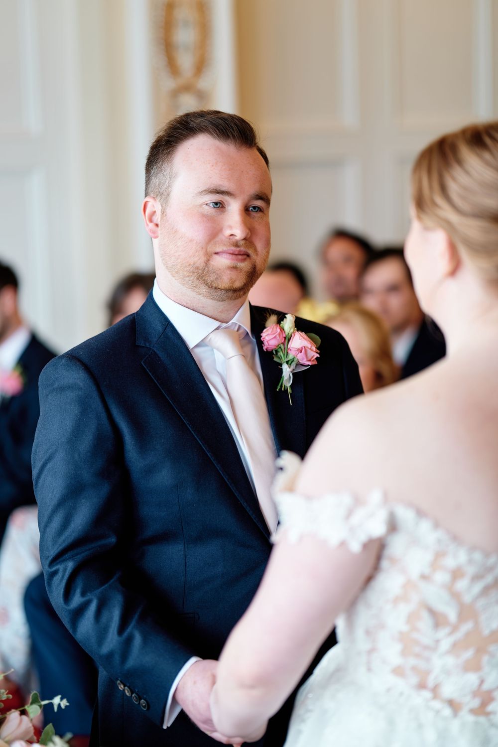 Groom in navy suit with pink boutonniere gazing at bride during wedding ceremony.
