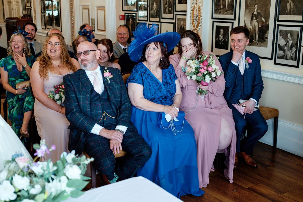 Wedding guests seated in rows, smiling emotionally, woman in blue hat holding bouquet, elegant venue with framed photos on walls.