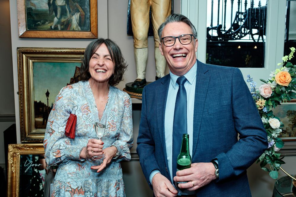 Two smiling guests holding drinks at an elegant indoor social gathering surrounded by artwork and floral arrangements.