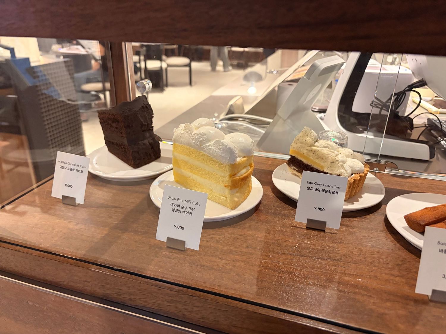 Glass display case in a cafe showcasing slices of chocolate cake, crepe cake, and pastries on white plates with price tags.