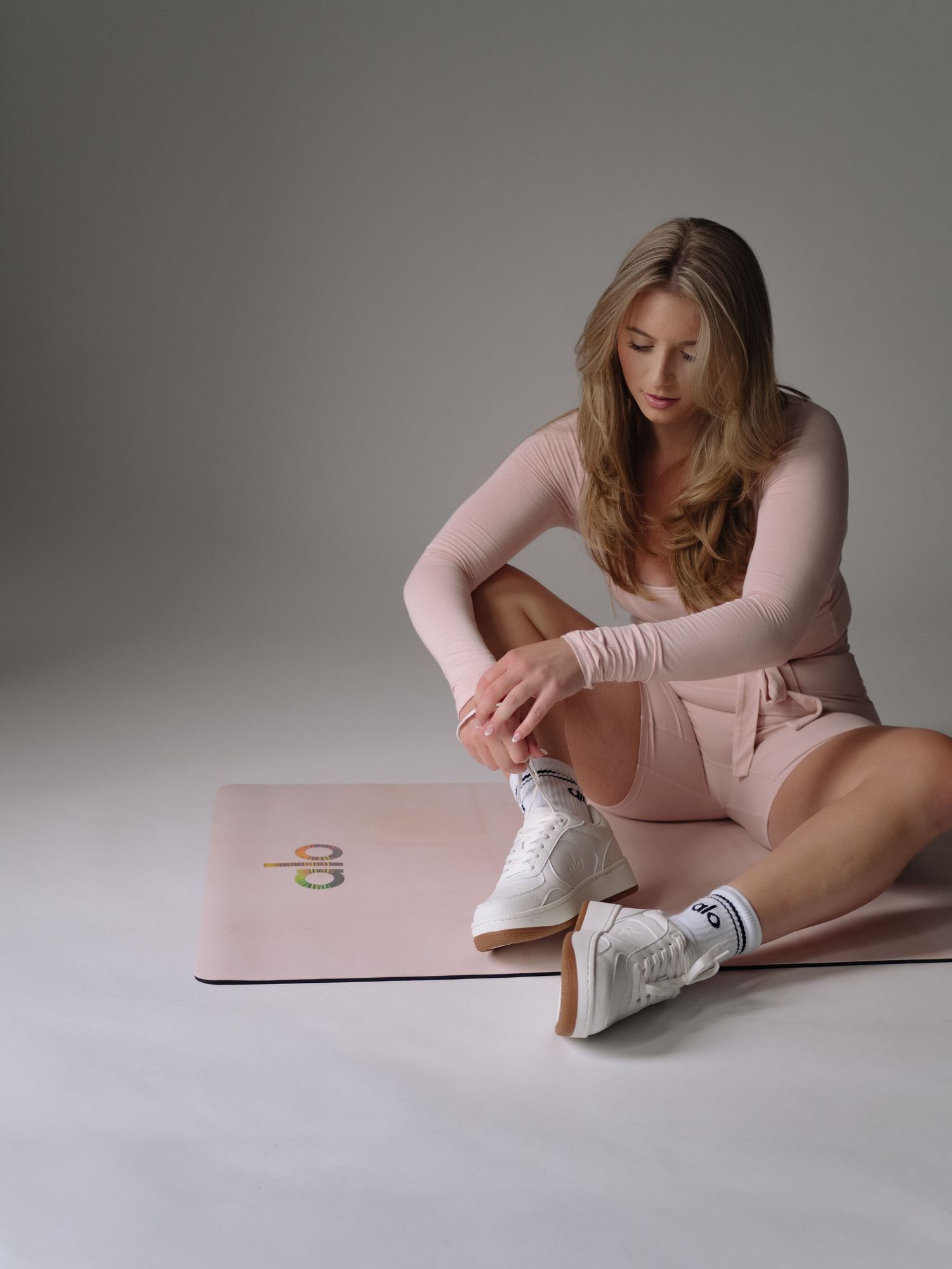 Woman in pink activewear sitting on a pink yoga mat wearing white sneakers against a gray background.