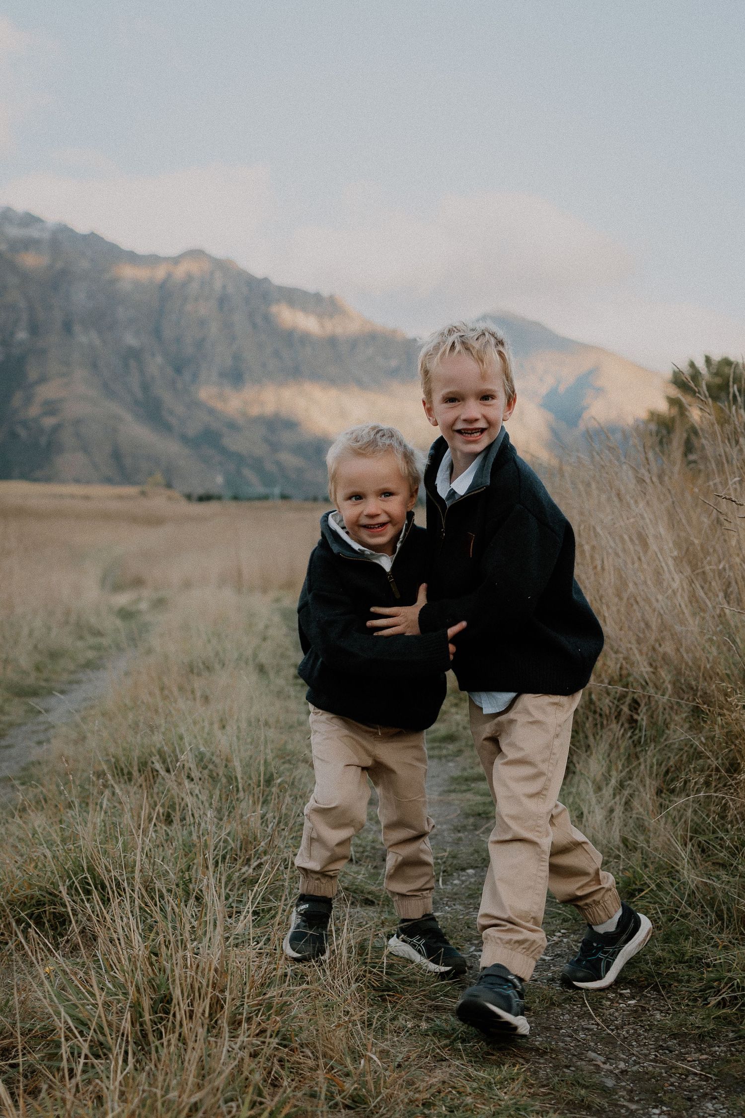 Family of five walking through mountain meadow, grandparents swinging child in air, snow-capped peaks backdrop.